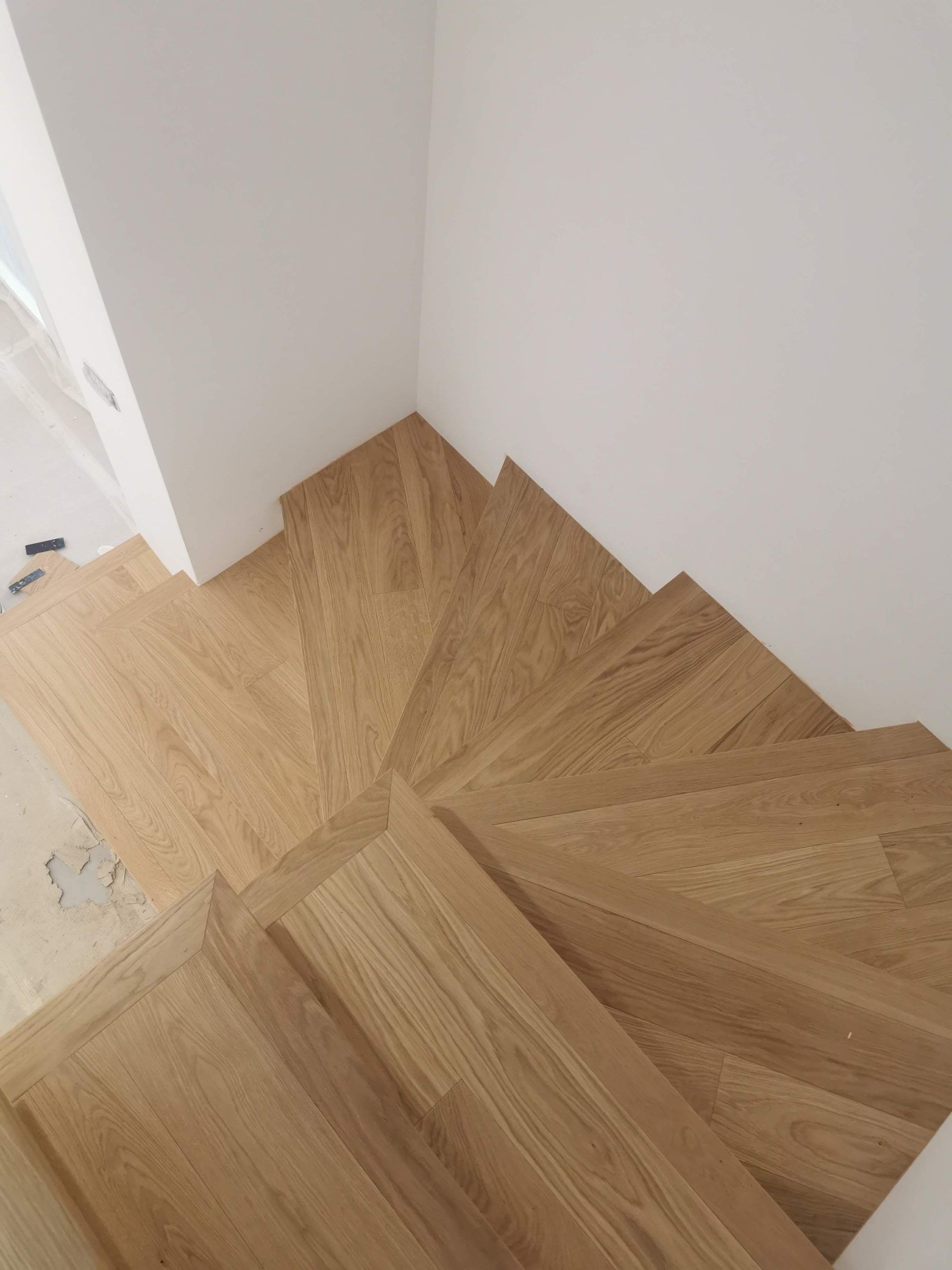 In un angolo di una stanza vuota, le assi di legno del pavimento posate a spina di pesce incontrano pareti bianche.