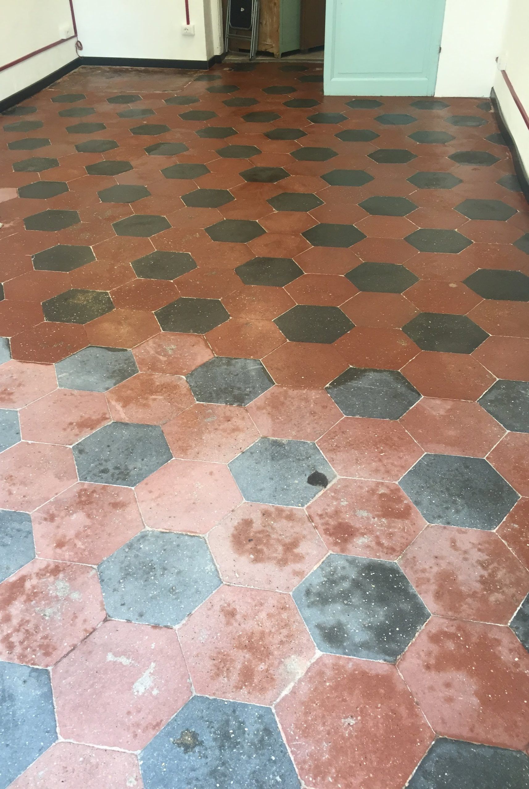 Un pavimento rosso consumato con piastrelle ottagonali scure in un corridoio o in una stanza, che conduce a una porta.