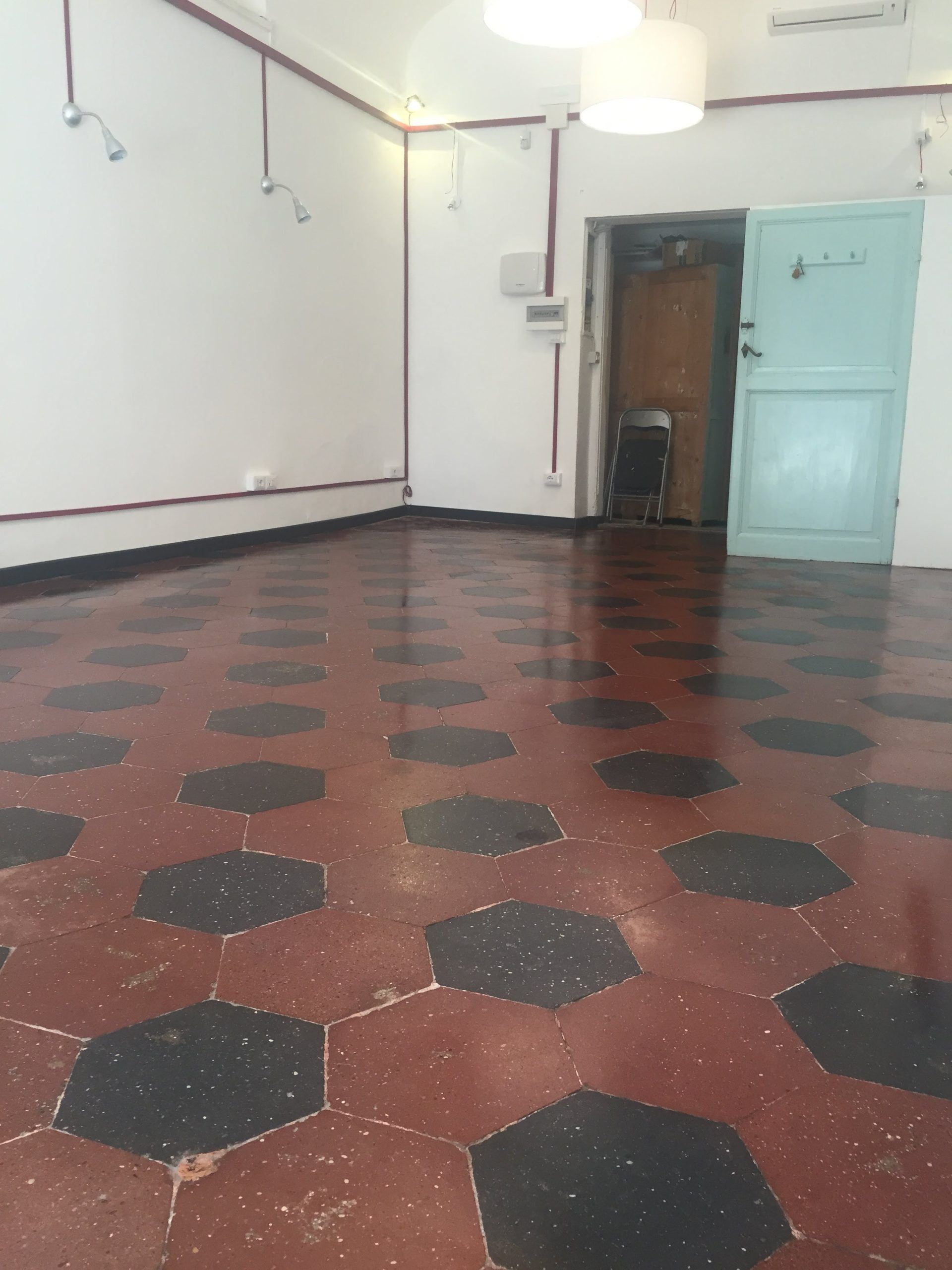 Stanza vuota con pavimento piastrellato con motivi rossi, pareti bianche e una porta color verde acqua sulla destra.