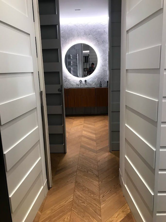 Cabina armadio con ripiani bianchi, pavimento in legno a spina di pesce e specchio rotondo illuminato sul fondo.