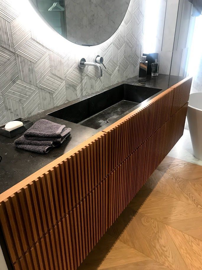 Mobile bagno moderno con piano nero, specchio rotondo, lavabo a parete e pannello frontale a doghe di legno.