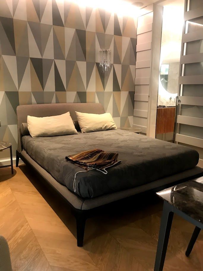 Camera da letto moderna con letto grigio, parete decorativa con motivi geometrici, pavimento in legno e ante dell'armadio a specchio.