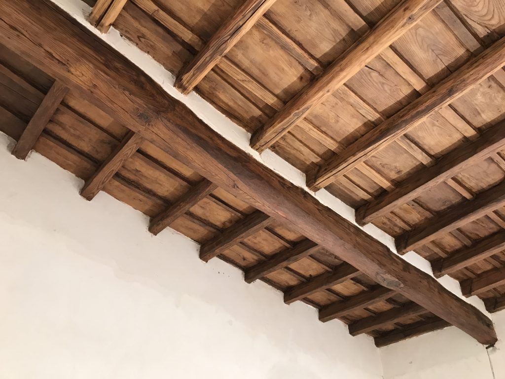 Soffitto con travi in legno e tegole in terracotta sopra una parete bianca.