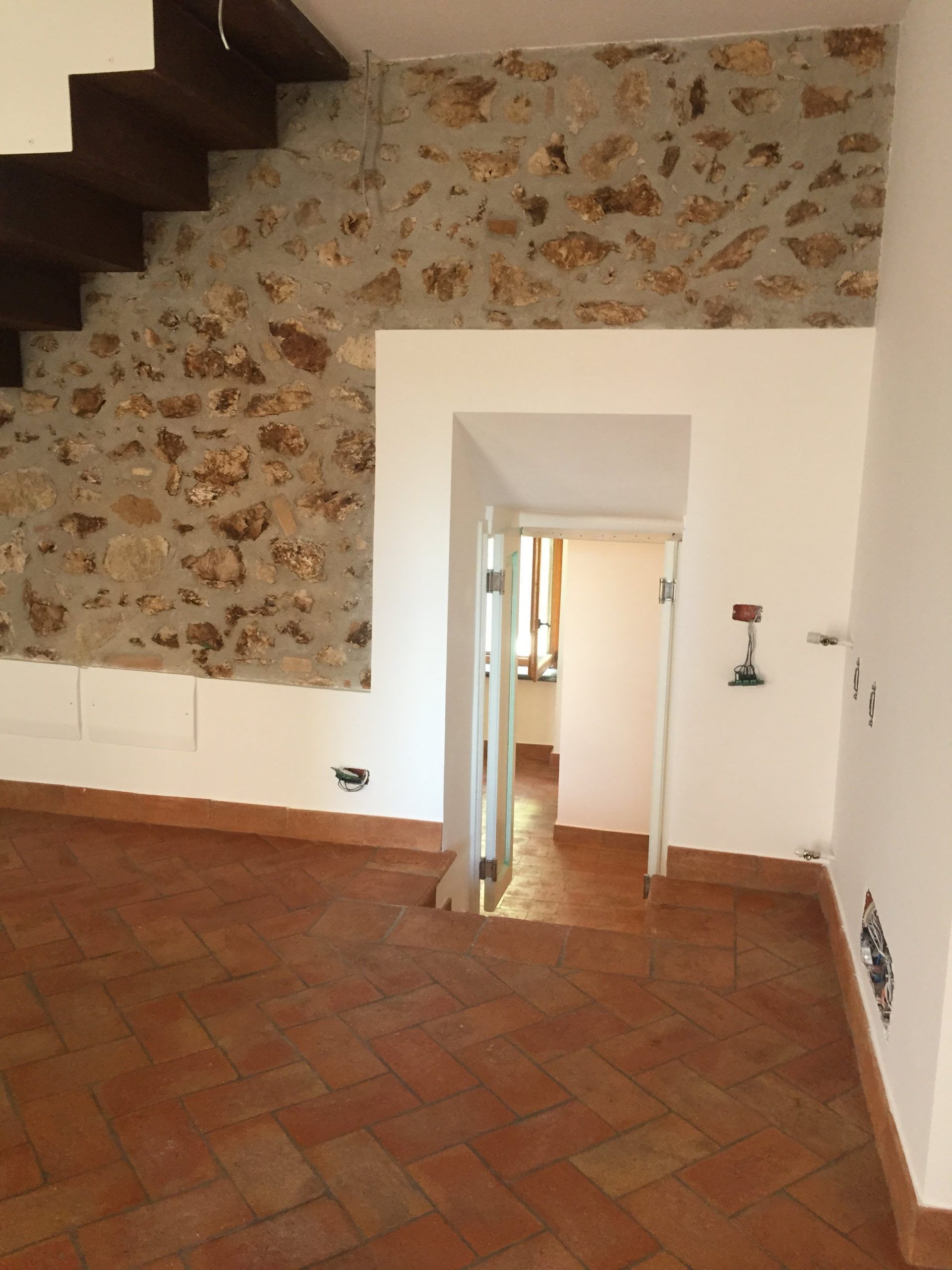 Stanza vuota con pavimento in piastrelle di terracotta, parete con rivestimento in pietra, scala e porta che conduce a un corridoio.