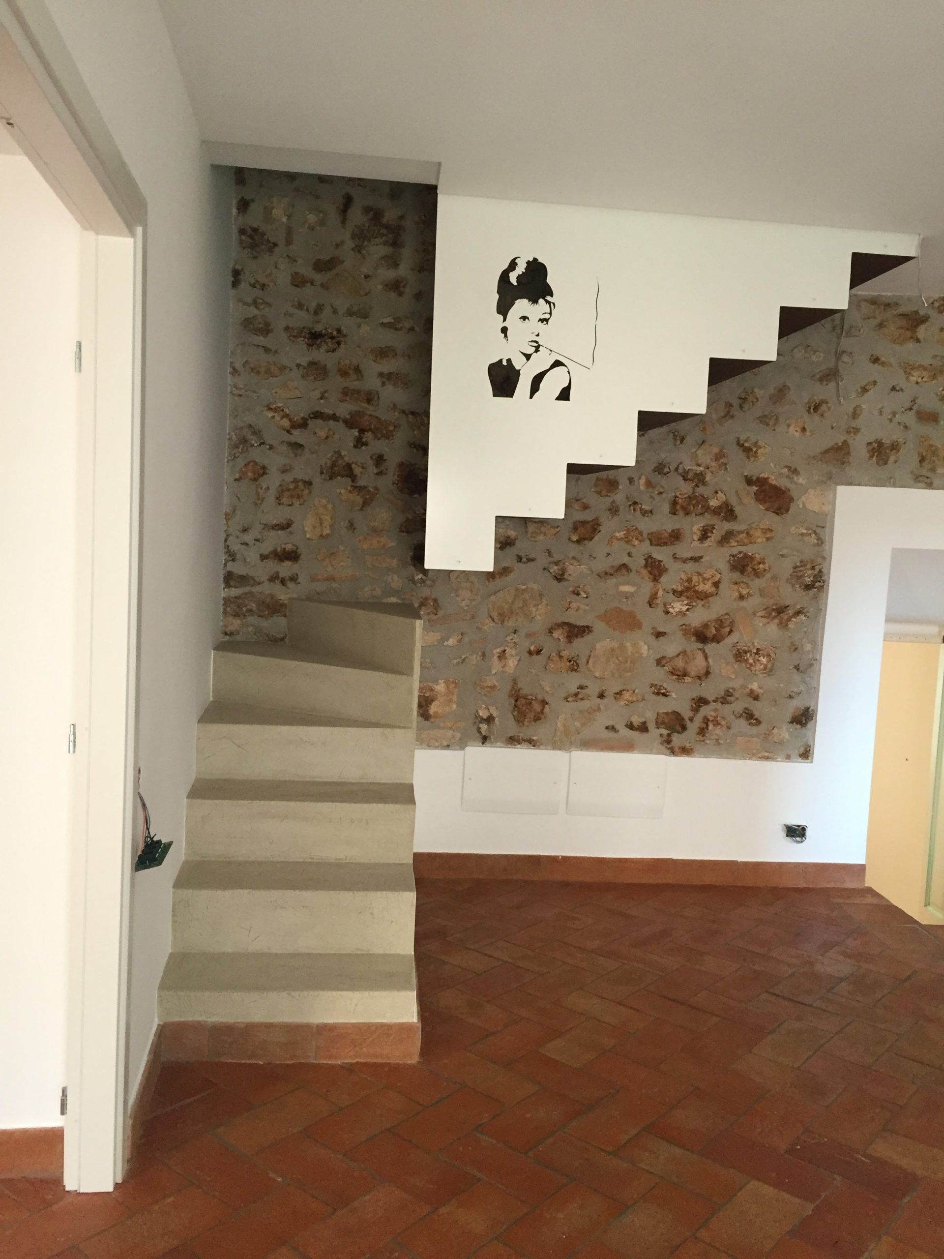 Interno di una stanza con parete in mattoni, scala, porta bianca e pavimento piastrellato di rosso.