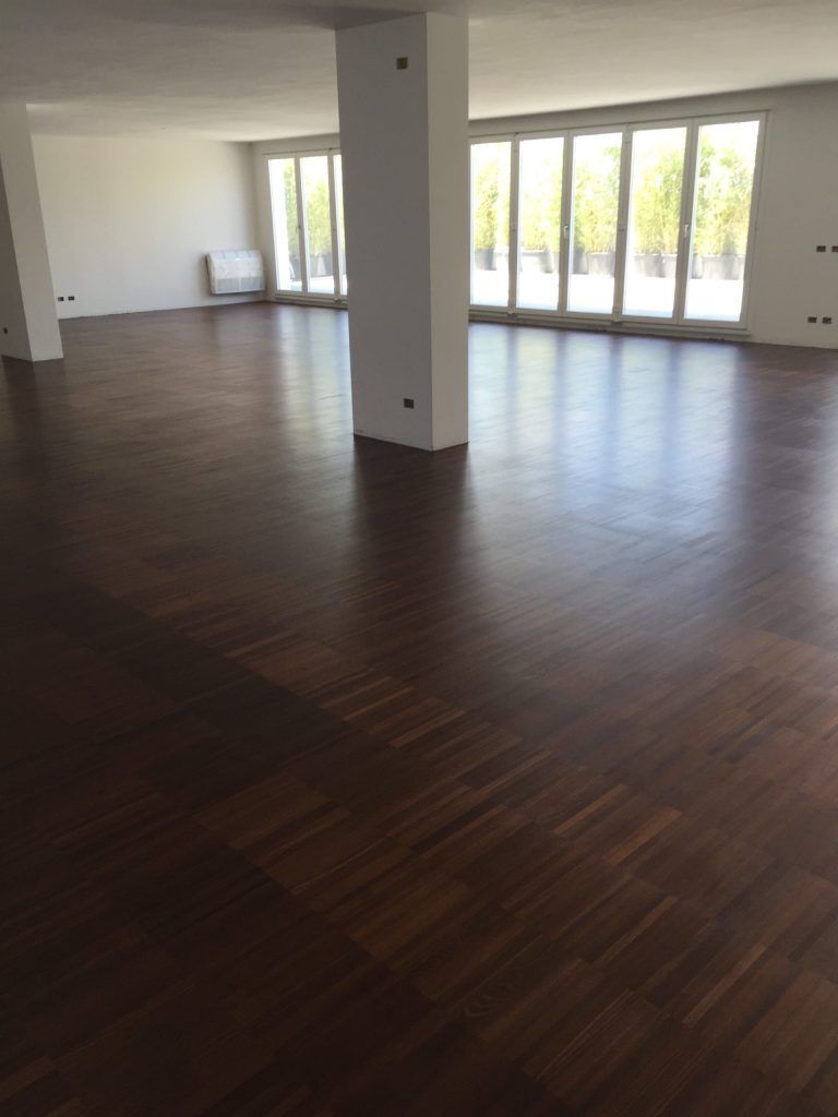 Stanza moderna vuota con pavimento in legno scuro, pareti bianche e ampie finestre che lasciano entrare la luce naturale.