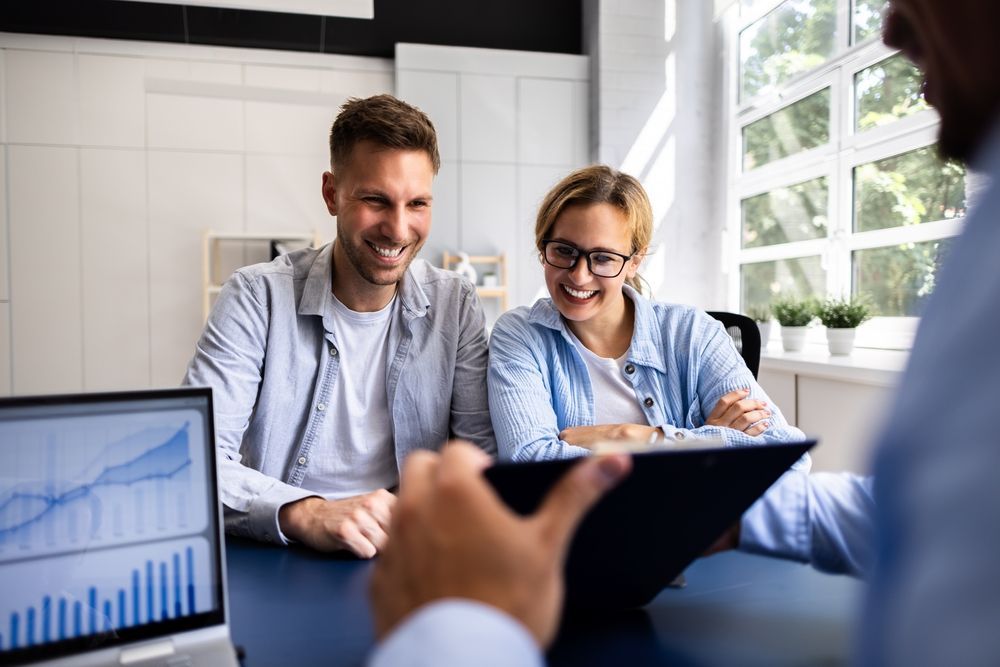 Two people smiling in a bright office meeting, with a chart on a laptop screen.