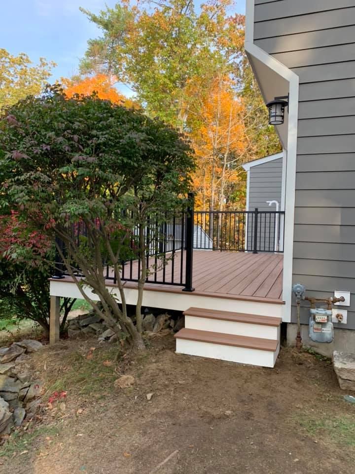 A wooden deck with black railings and a two-step staircase attached to the side of a gray-sided house near autumn trees.