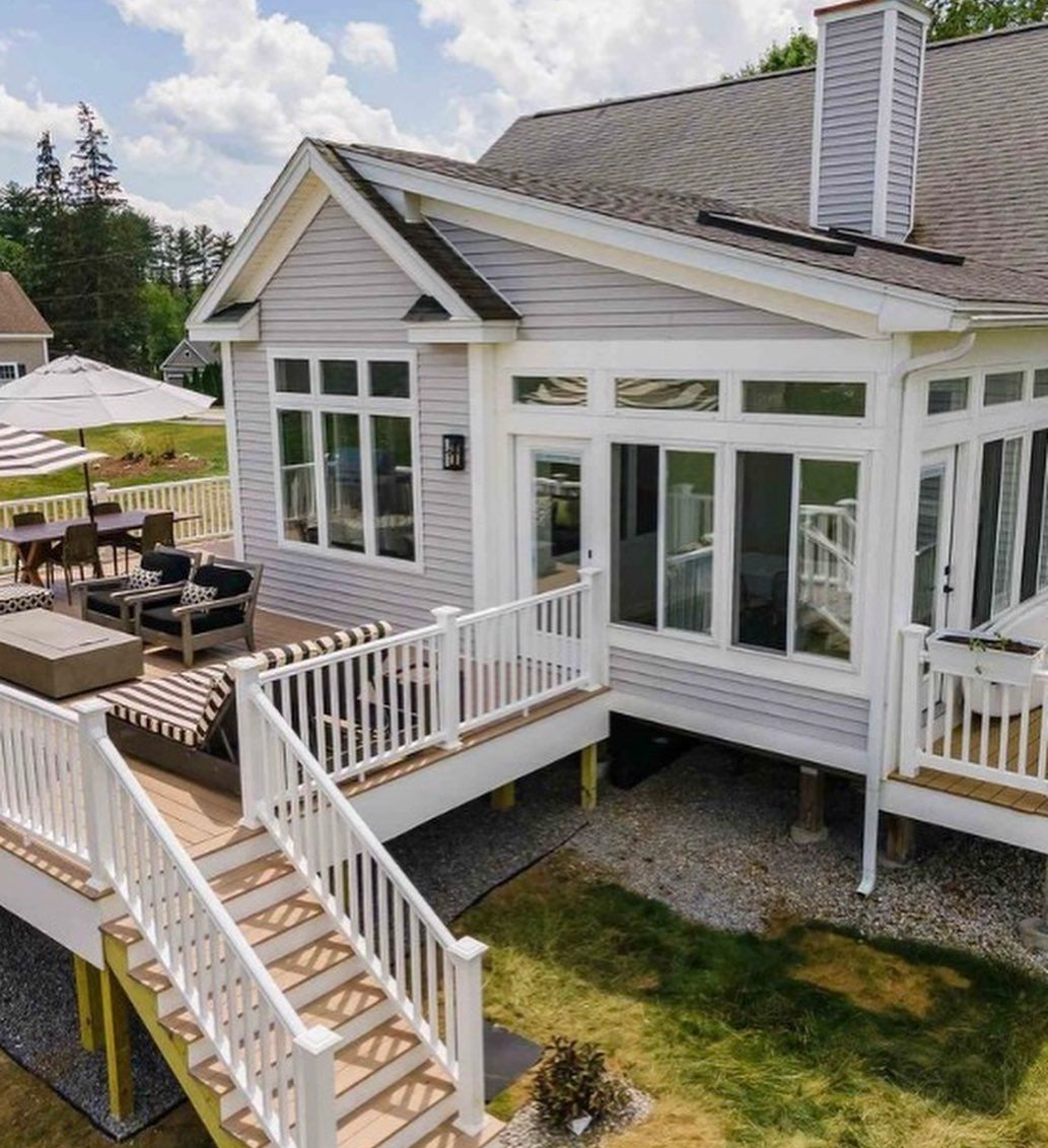 Elevated wooden deck with stairs and patio furniture attached to a light gray house with large windows.