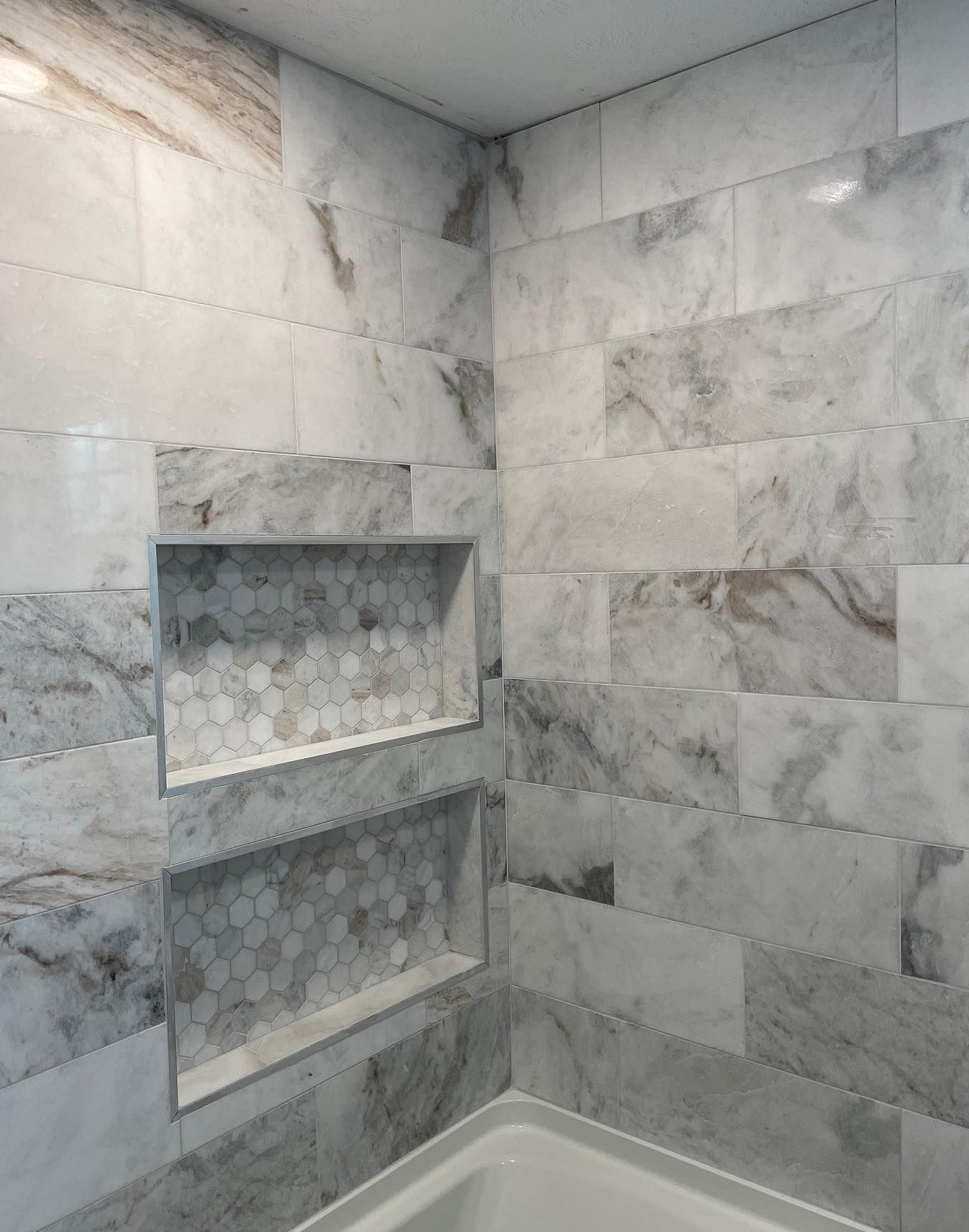 A corner of a tiled shower with two built-in storage niches featuring a gray and white marble pattern throughout.