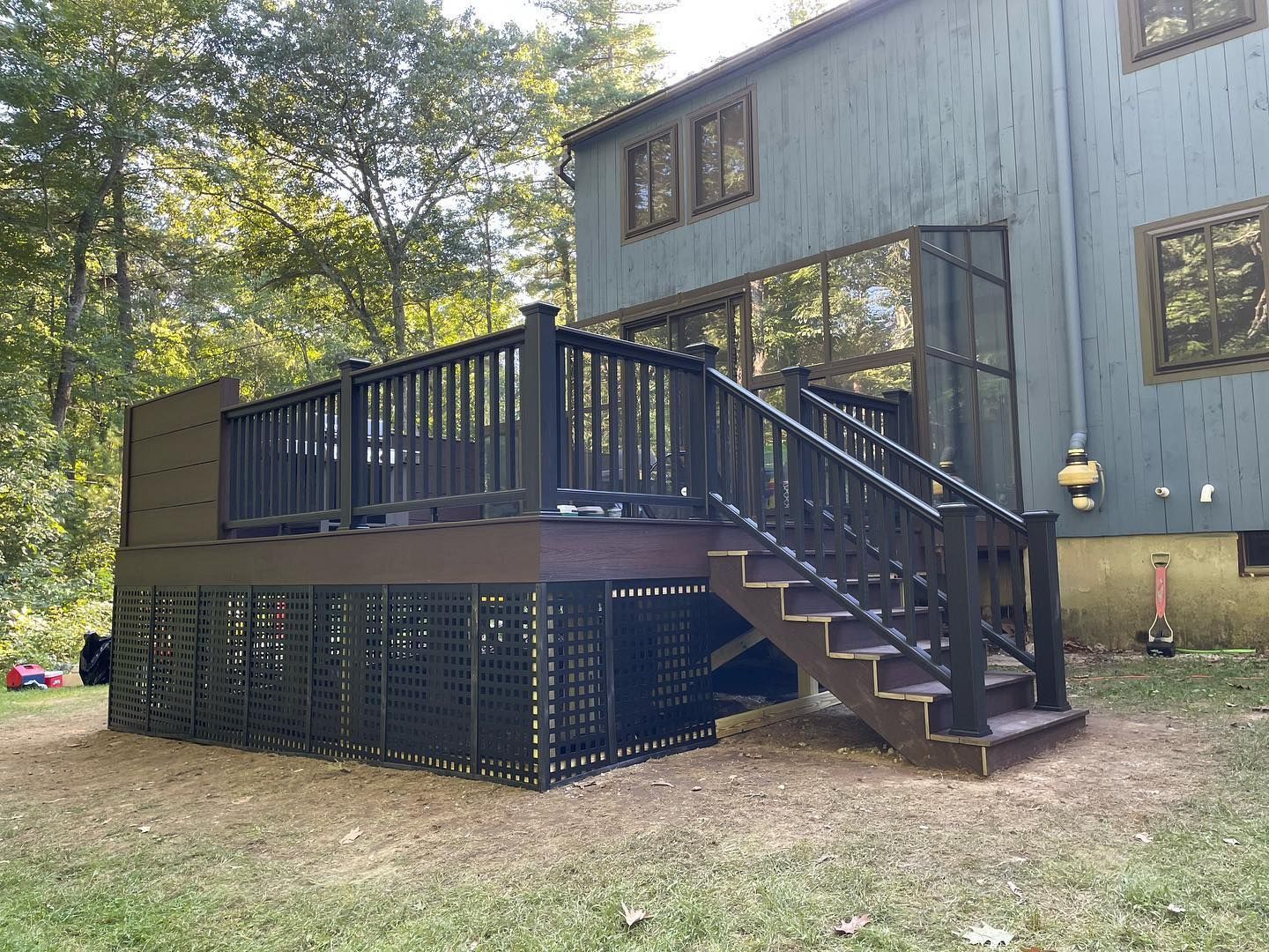 A dark brown wooden deck with black railings and stairs, featuring latticework skirting, attached to a gray house.