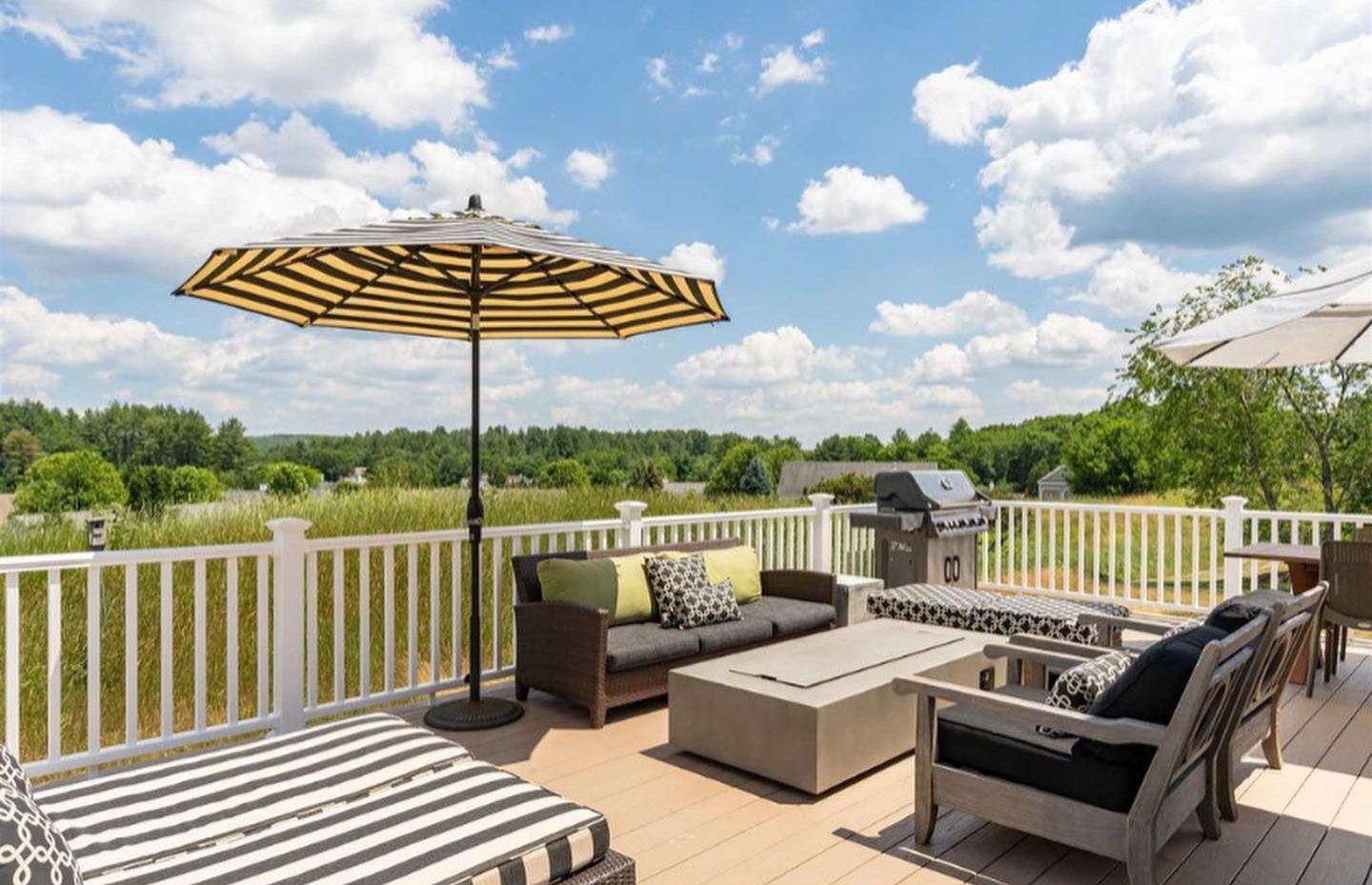 A sunny outdoor deck with wicker furniture, a rectangular fire pit, striped umbrellas, and a grill overlooking a field.