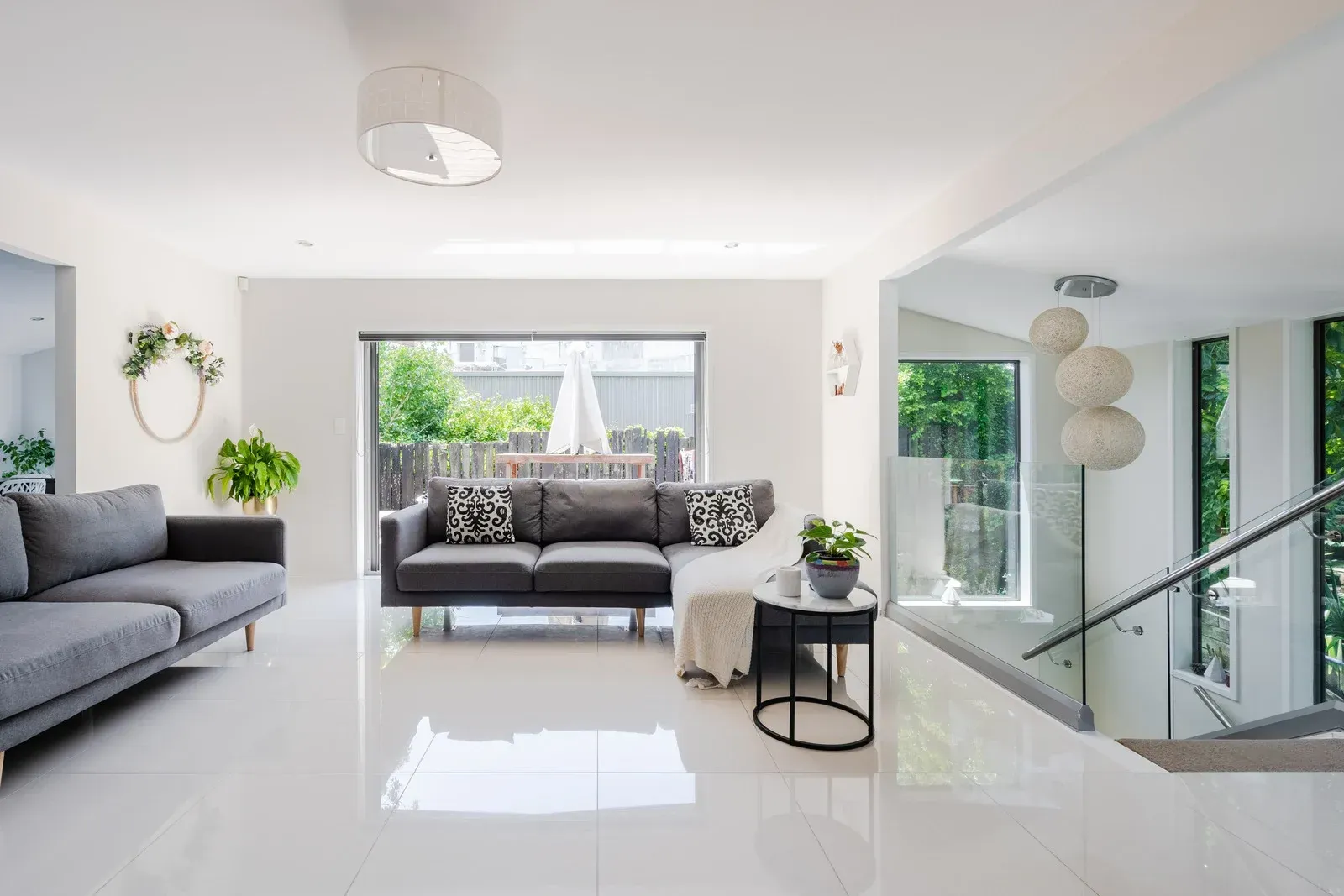 Modern living room with a gray sectional, white tiled floors, large windows, and a glass-paneled staircase.