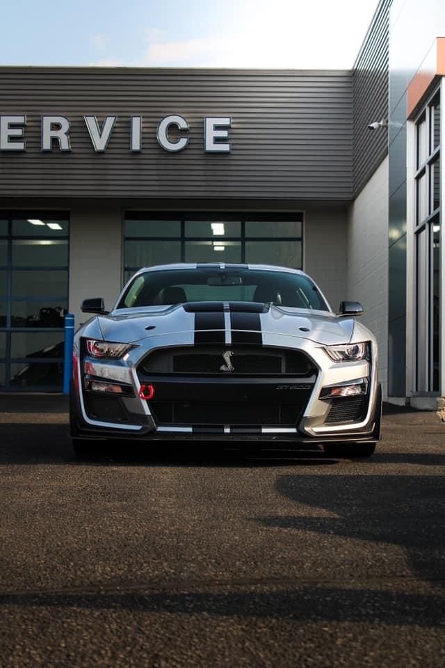 Silver Ford Mustang Shelby in front of a service center, with black stripes, a black grille, and the word 
