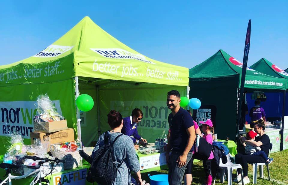 Outdoor fair booth with lime-green tents, people chatting, and children seated nearby on a sunny day