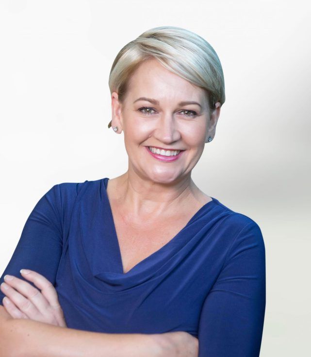 Smiling blonde woman in a blue top with arms crossed against a light background
