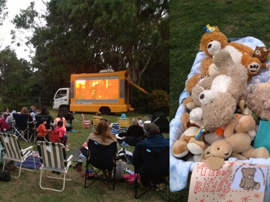 Outdoor gathering with people seated on chairs watching a screen beside a large teddy bear display.