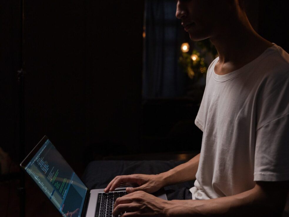 A man is typing on a laptop computer in a dark room.