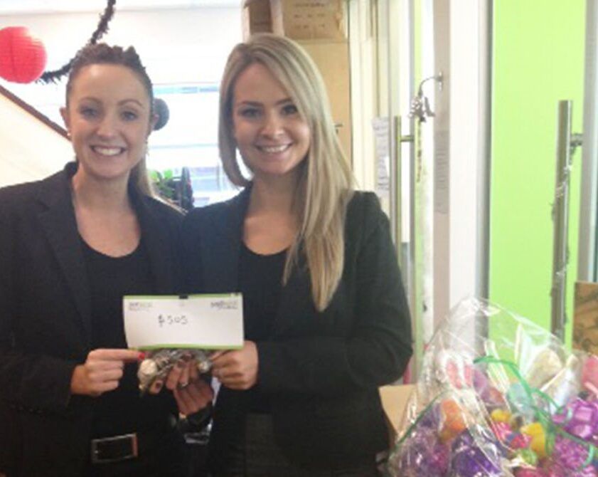 Two women in black outfits smiling and holding a check in a bright office, with colorful gifts beside them