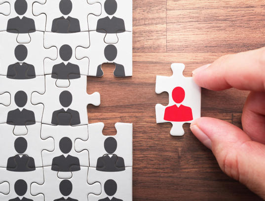 Hand placing a red puzzle piece into a group of gray puzzle pieces on wood background
