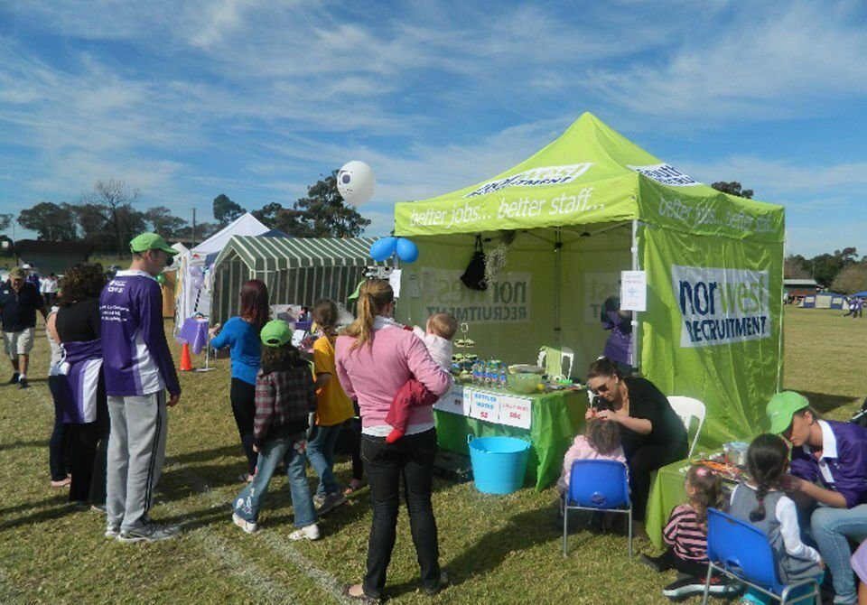 People gathered at a bright green booth outdoors on a sunny day, with a banner and seated staff.