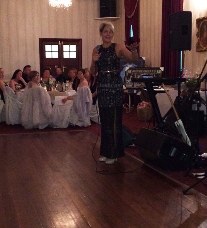 Woman speaking at a banquet hall microphone beside a keyboard, with seated guests in white dresses behind her.