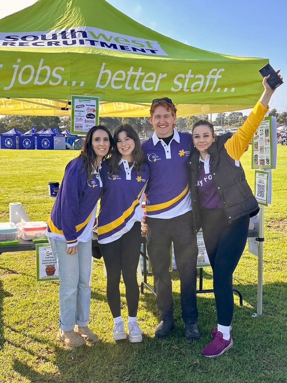 Four people posing under a green tent with “jobs... better staff...” banner at an outdoor event
