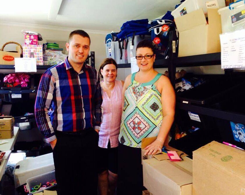 Three people smiling in a cluttered storage room with shelves, boxes, and supplies