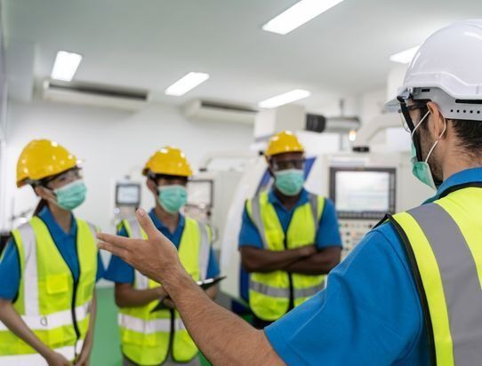 Workers in safety gear listening to a supervisor in a factory break room