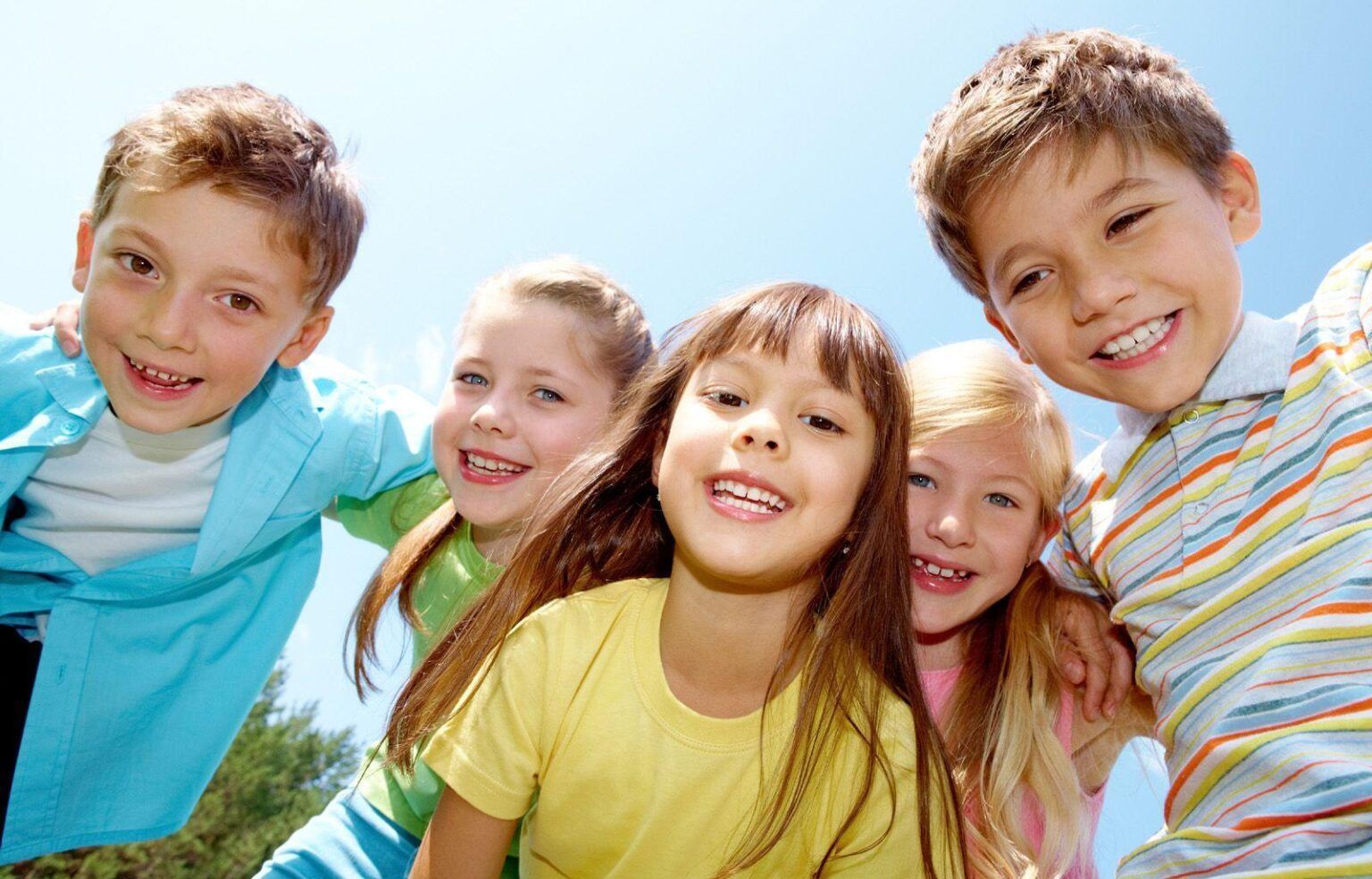 Five smiling children leaning together outdoors under a blue sky