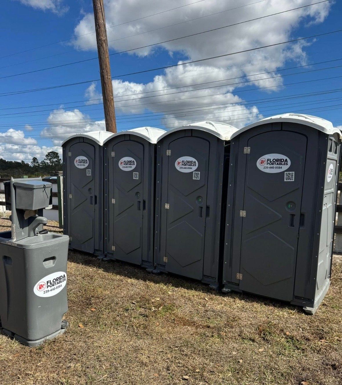 Four gray portable toilets and a handwashing station outdoors under a blue sky.