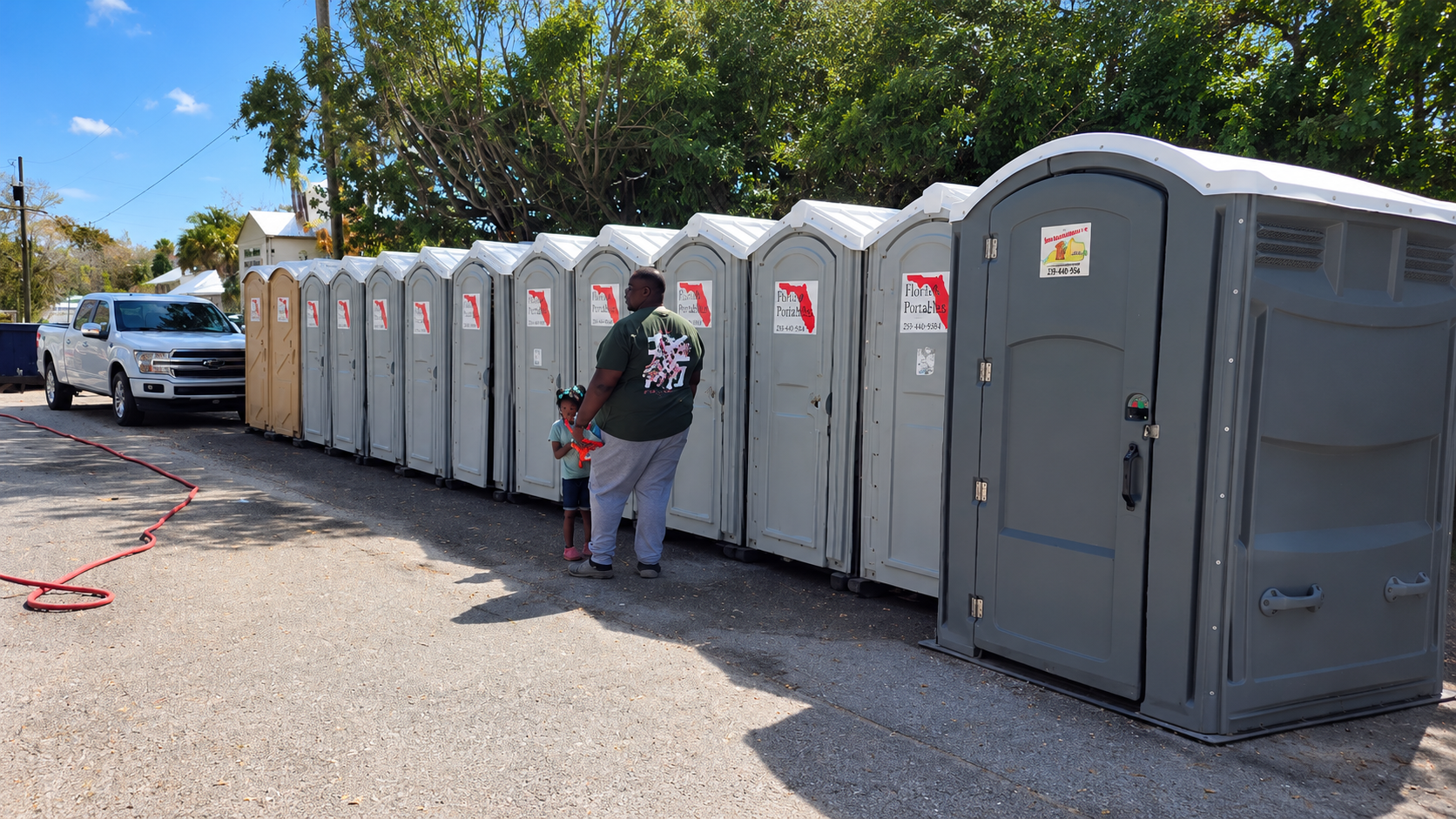 Row of portable toilets beside a parked truck, with a person standing in front on a gravel lot.