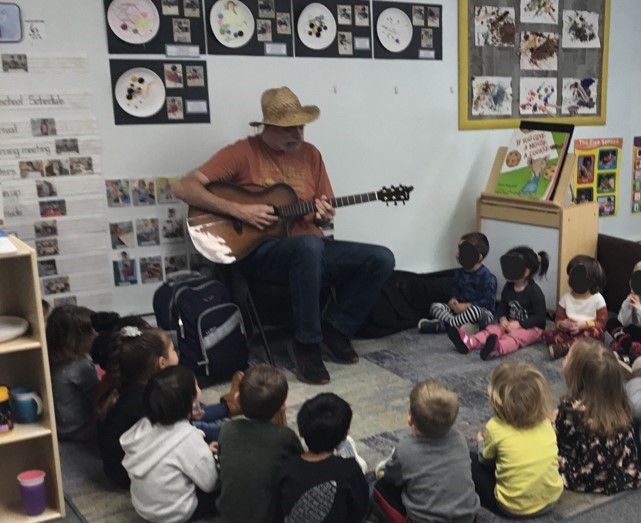 Man playing guitar, singing to a group of children seated on the floor in a classroom.