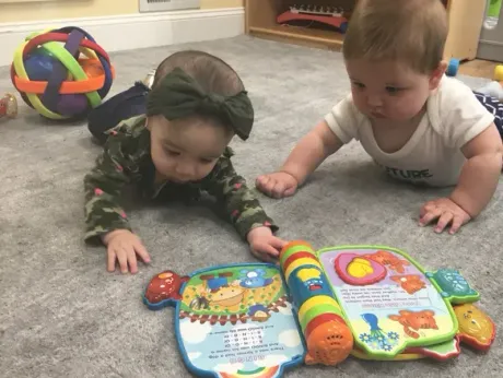 Two babies on carpet, reaching for a colorful toy book. One wears a bow, the other a white onesie.