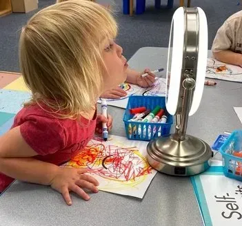 Young child looks at themselves in a lighted mirror while coloring a picture with markers at a desk.