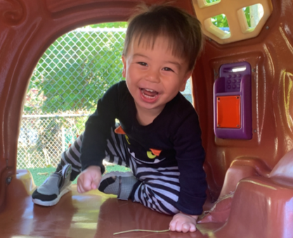 Young child smiles, crawling in a playhouse, wearing black long-sleeve shirt and striped pants.