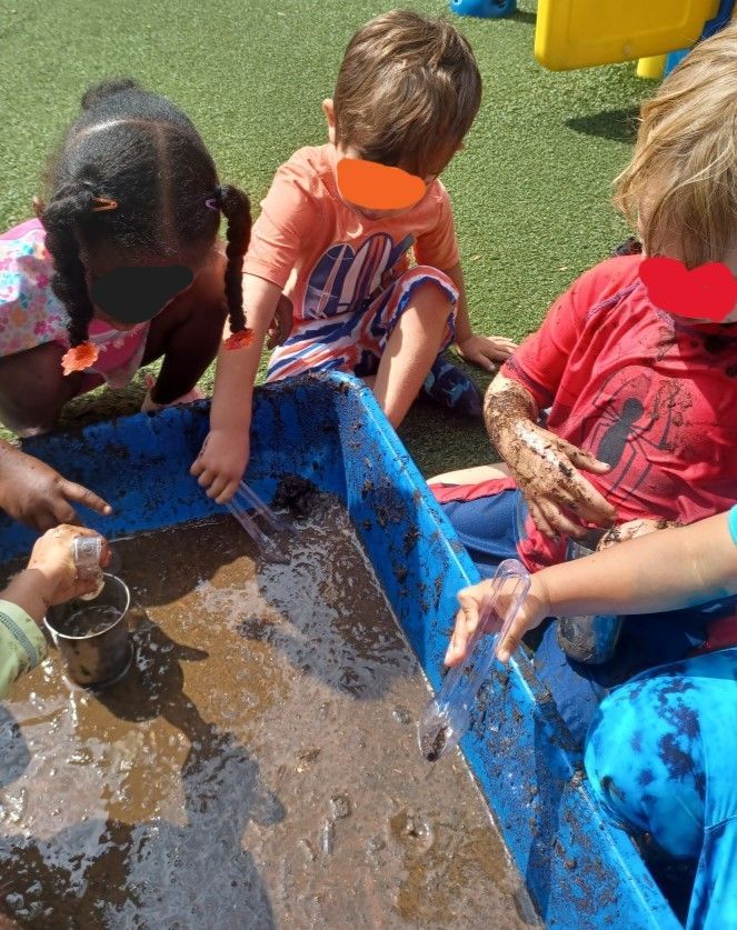Children playing with mud in a blue bin outdoors. Hands covered in mud, some pointing and holding tools.
