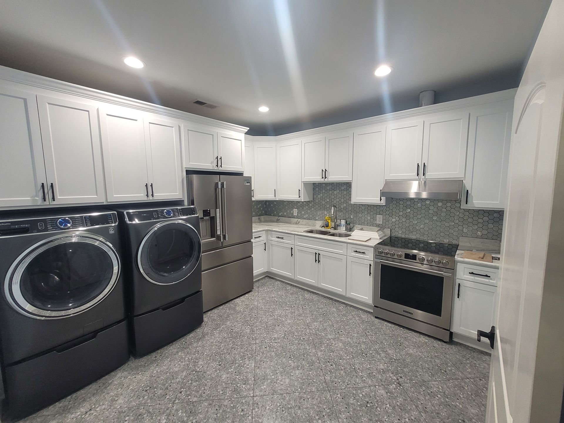 Laundry machines and kitchen, white cabinets, stainless steel appliances. Grey speckled floor.