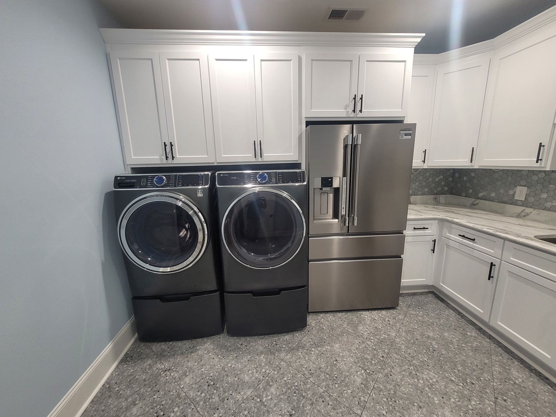 Laundry room with gray washer/dryer, stainless fridge, white cabinets, and speckled gray floor.