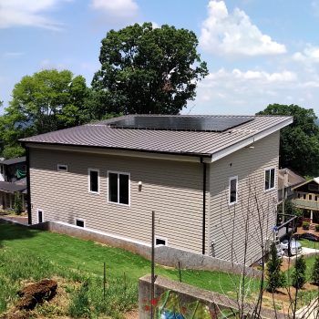 Modern tan house with a dark gray roof, solar panels, and multiple windows, set on a grassy hill.