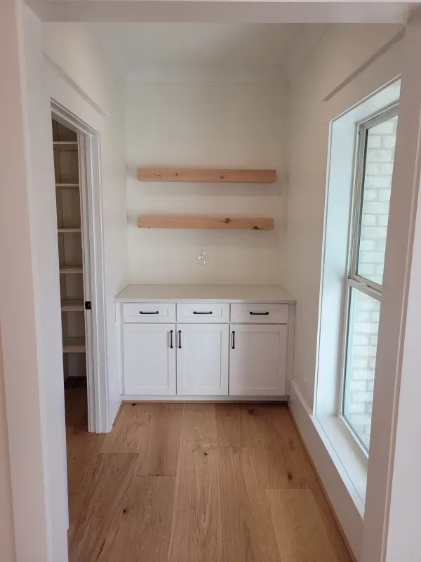 White built-in cabinets with floating shelves and a window in a neutral room, wooden floor.