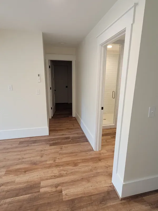 A hallway with wood-look floors, white walls, and doorways leading to a bathroom and another room.