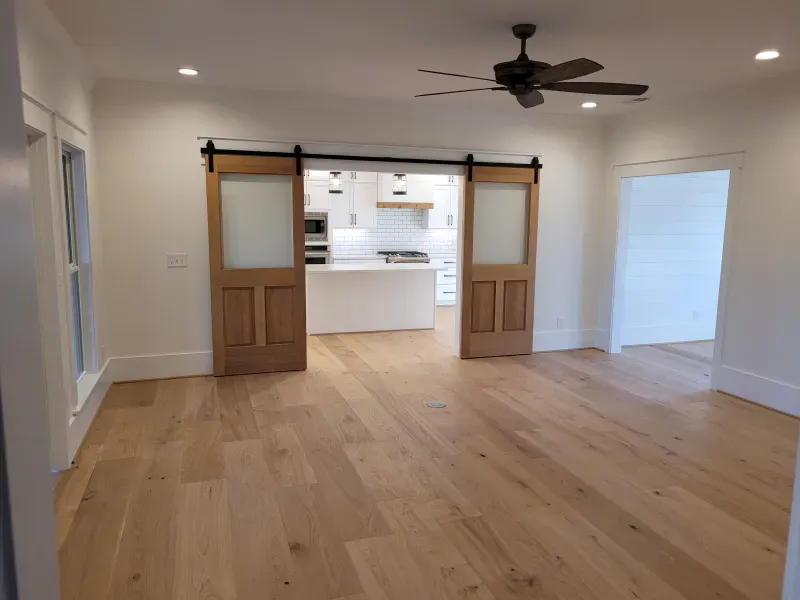 Empty room with light wood floor, two barn doors, and a view into a white kitchen.