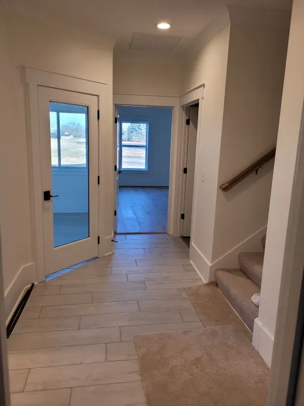 Hallway with light-colored tile floor, open doorway to a room with a window, and staircase with a handrail.