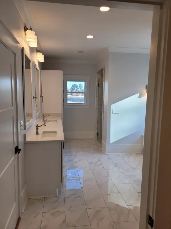 A white bathroom with a long vanity, marble floors, and a window at the end.