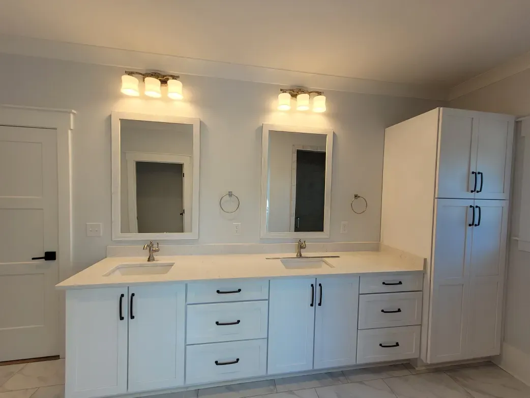 Modern white bathroom with double vanity, mirrors, and cabinet; gold accents, black hardware.
