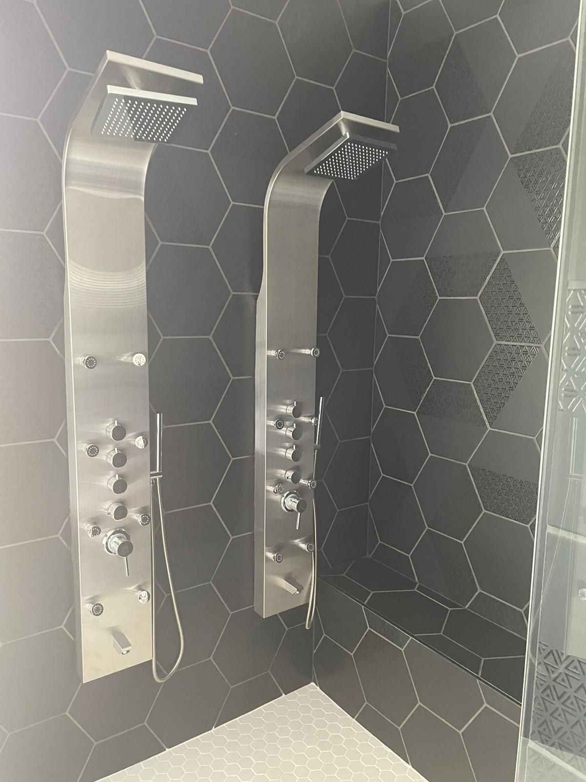 Two stainless steel shower panels with multiple jets and shower heads, installed on a black hexagon tile wall.