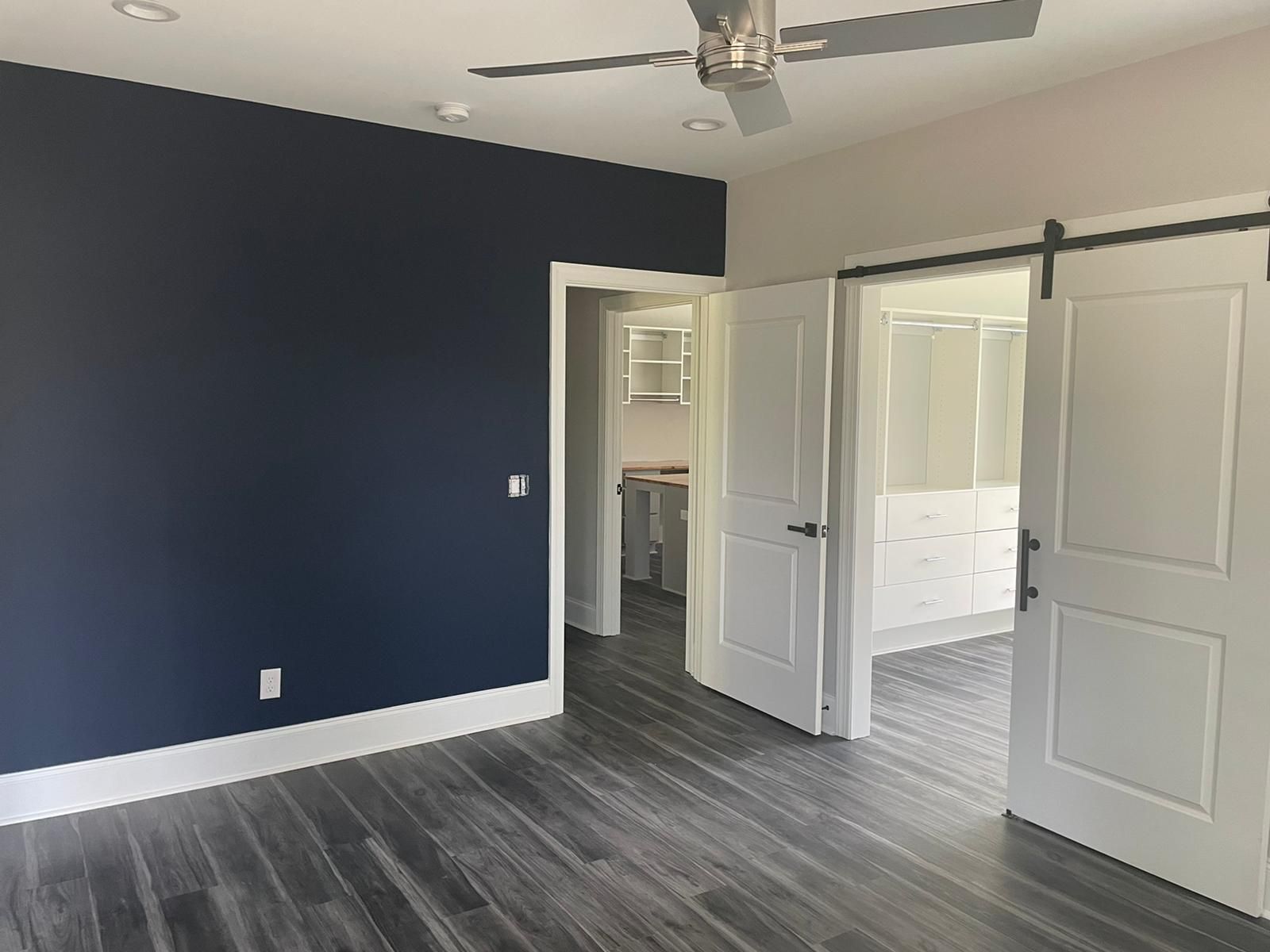 A bedroom with dark blue wall, white doors, and grey floors. Door leads to a bathroom and kitchen.