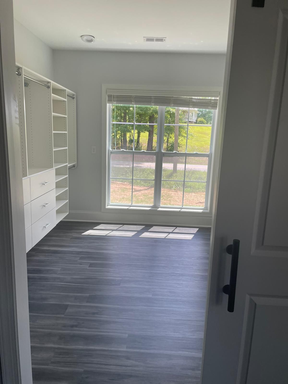 Walk-in closet with white shelving, dark wood-look floor, and large window overlooking trees.