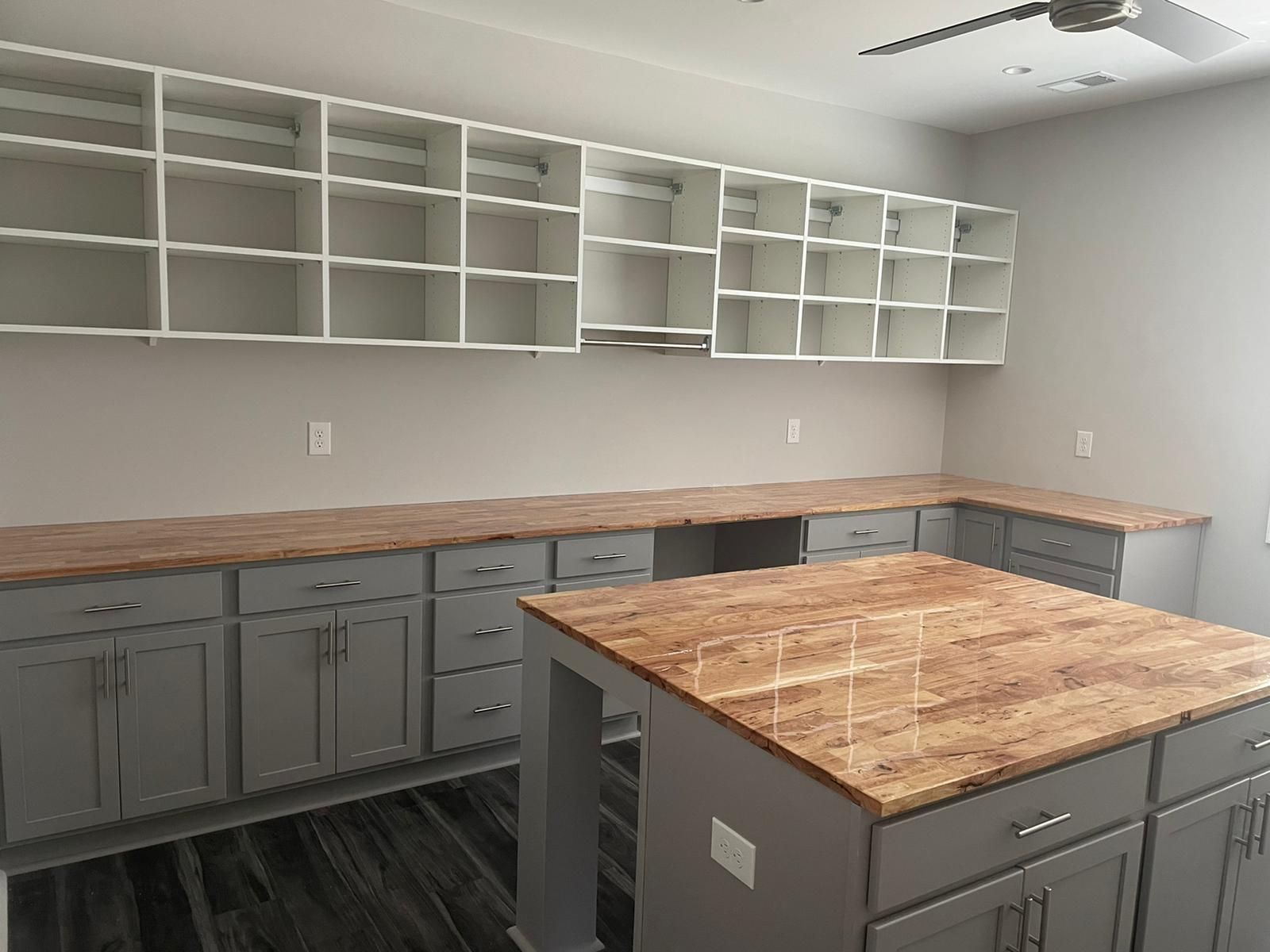 Gray cabinetry with butcher block countertops and upper shelving in a room; a ceiling fan.